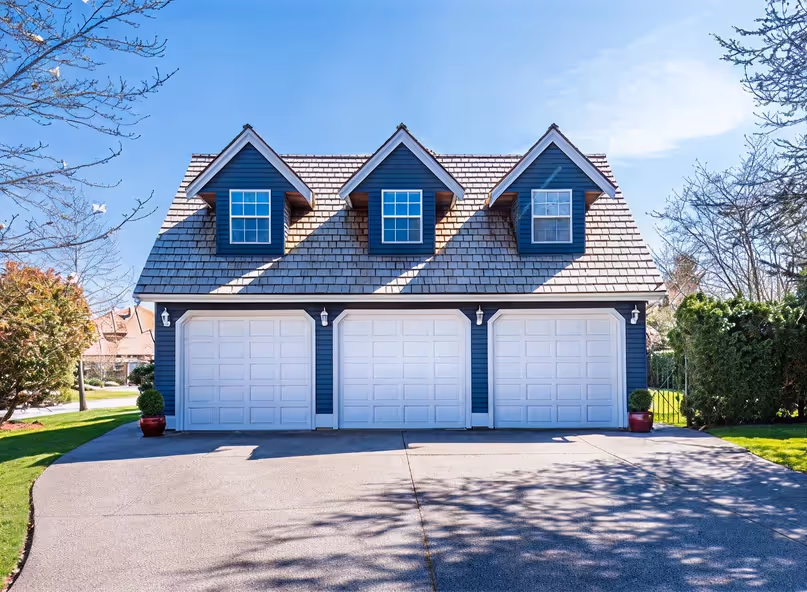 Blue garage building with three white doors and three dormer windows on a clear sunny day.