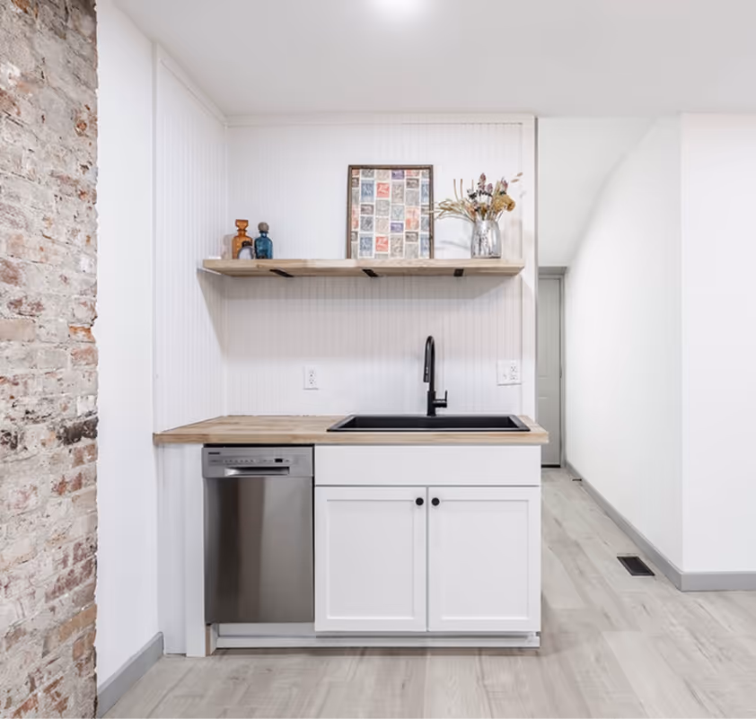 Small modern kitchen corner with white cabinets, wood countertop, black sink and faucet, stainless steel dishwasher, and a wooden floating shelf with decorative items.