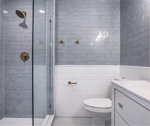 Modern bathroom with a glass-enclosed shower featuring brass fixtures, white toilet, and white vanity with gray tiled walls.