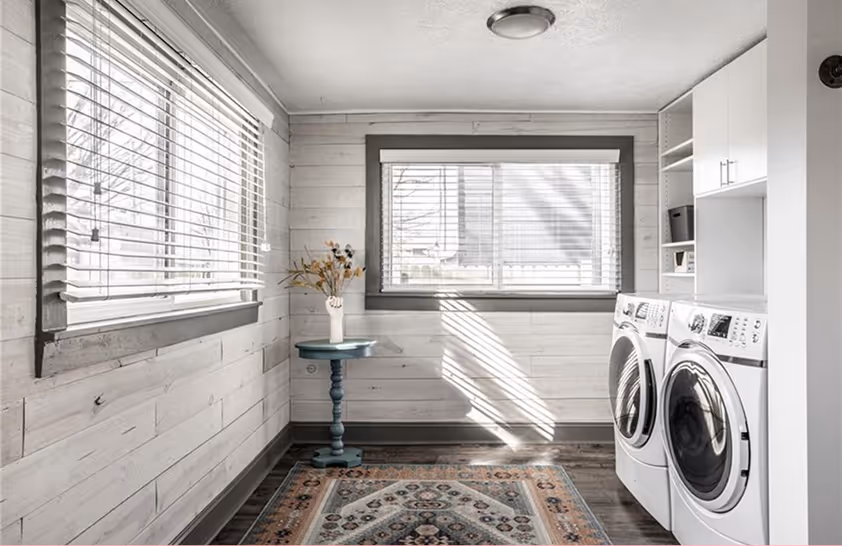 Bright laundry room with white walls, large windows with blinds, a patterned rug, a small blue side table with a vase of flowers, and a white washer and dryer set beside white storage shelves.