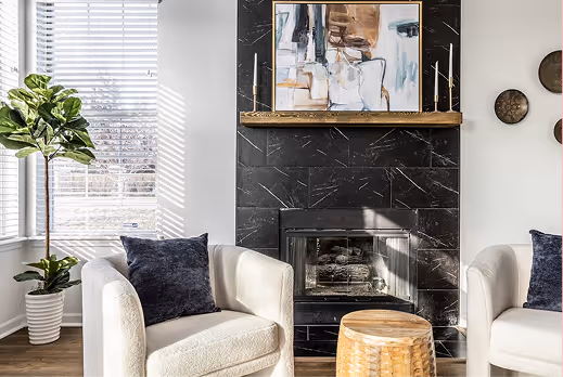 Bright living room with two white armchairs with dark pillows, a wooden side table, a black marble fireplace, framed abstract art, and leafy plants near a window with blinds.