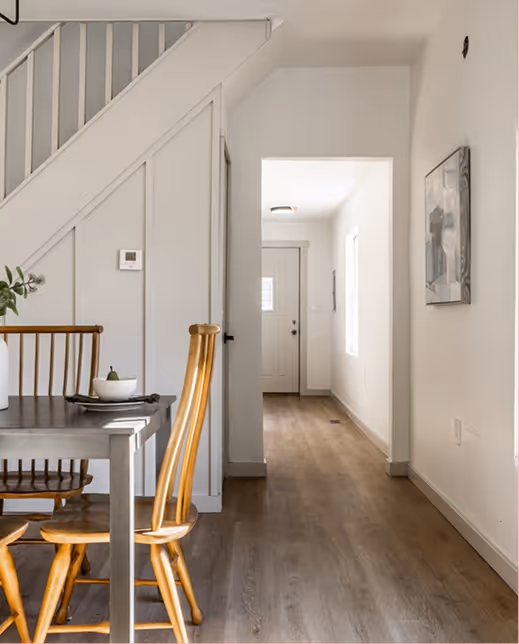 Bright hallway with wooden floor leading to front door next to a dining table and wooden chairs beneath a staircase.
