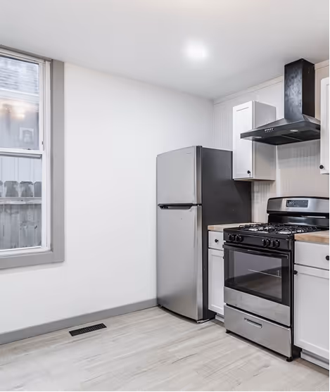 Modern kitchen corner with stainless steel refrigerator and gas stove under black range hood, white cabinets, and light wood floor.