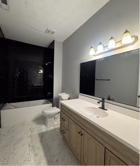 Modern bathroom with white marble floor, wooden vanity with white countertop and black faucet, large mirror, and a bathtub with black tiled walls.