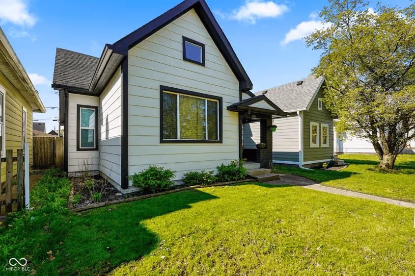 Small light-colored house with black trim, front porch, lush green lawn, and a large tree on the right side.