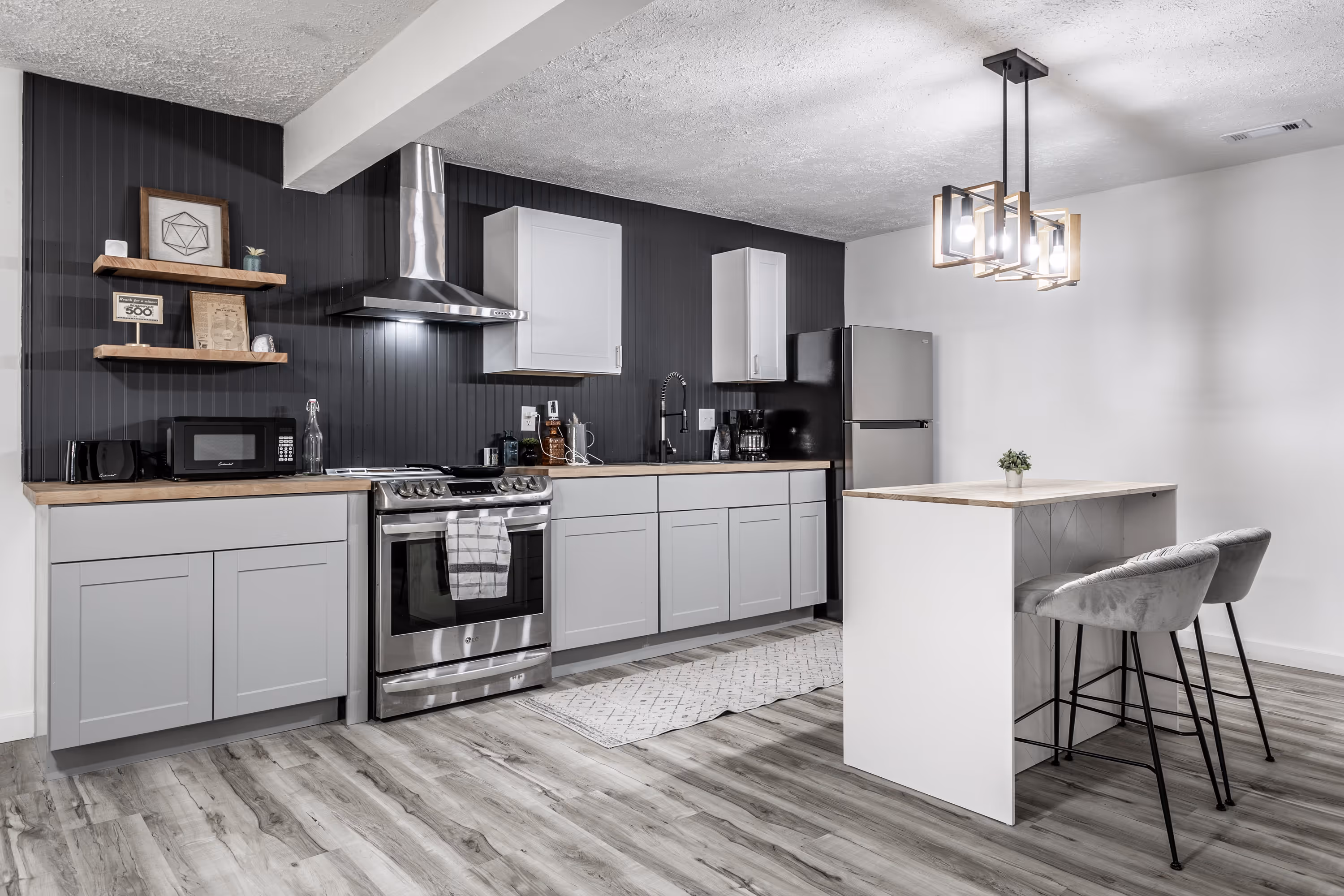 Modern kitchen with light gray cabinets, black backsplash, stainless steel appliances, wooden countertops, and a small island with two gray bar stools under a geometric pendant light.