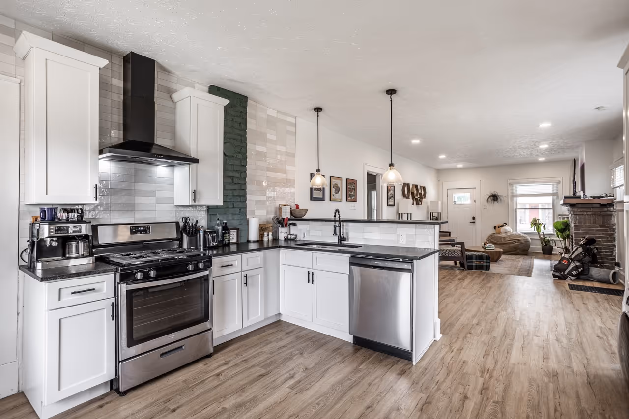 Modern kitchen with white cabinets, stainless steel appliances, black countertop, two pendant lights, and open view into living area with wood flooring.