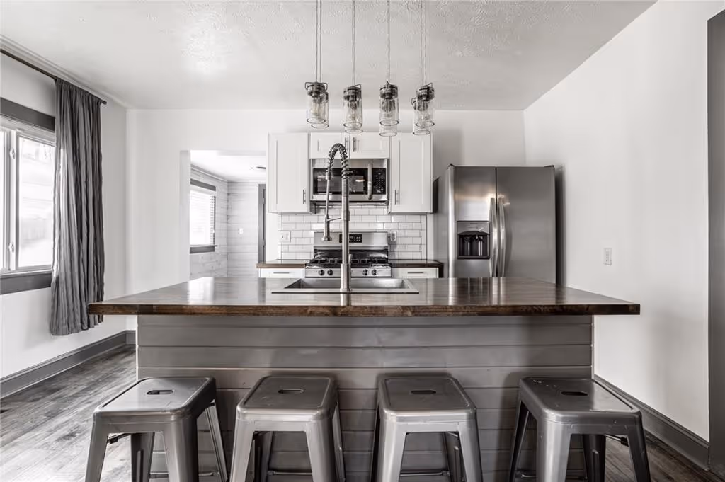 Modern kitchen with a large wooden island, four metal stools, stainless steel appliances, and hanging pendant lights.