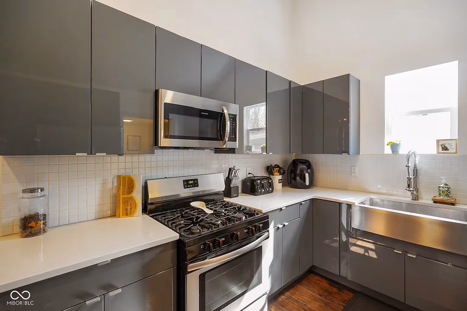 Modern kitchen with gray glossy cabinets, stainless steel stove and microwave, white countertops, and a large stainless steel sink under a bright window.