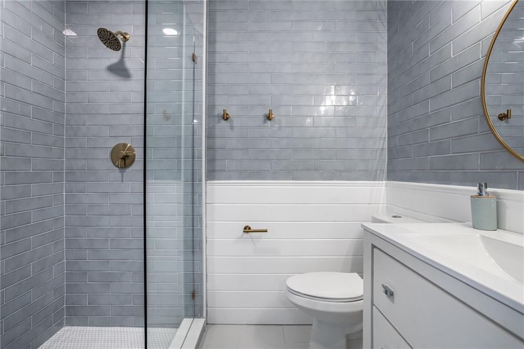 Modern bathroom with glass-enclosed shower featuring gray subway tiles, white toilet, white vanity with sink, round mirror, and gold fixtures.