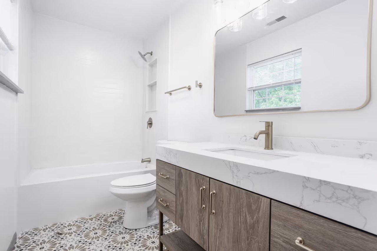 Modern bathroom with patterned tile floor, white bathtub, toilet, and wooden vanity with marble countertop and brass faucet beneath a large mirror.