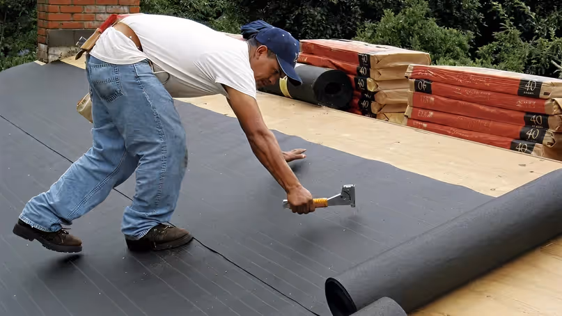 Man in blue jeans and white shirt installing black roofing underlayment on a wooden roof deck.