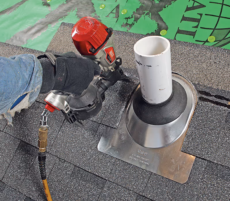 Person wearing gloves using a pneumatic nail gun to install shingles near a vent pipe on a roof.