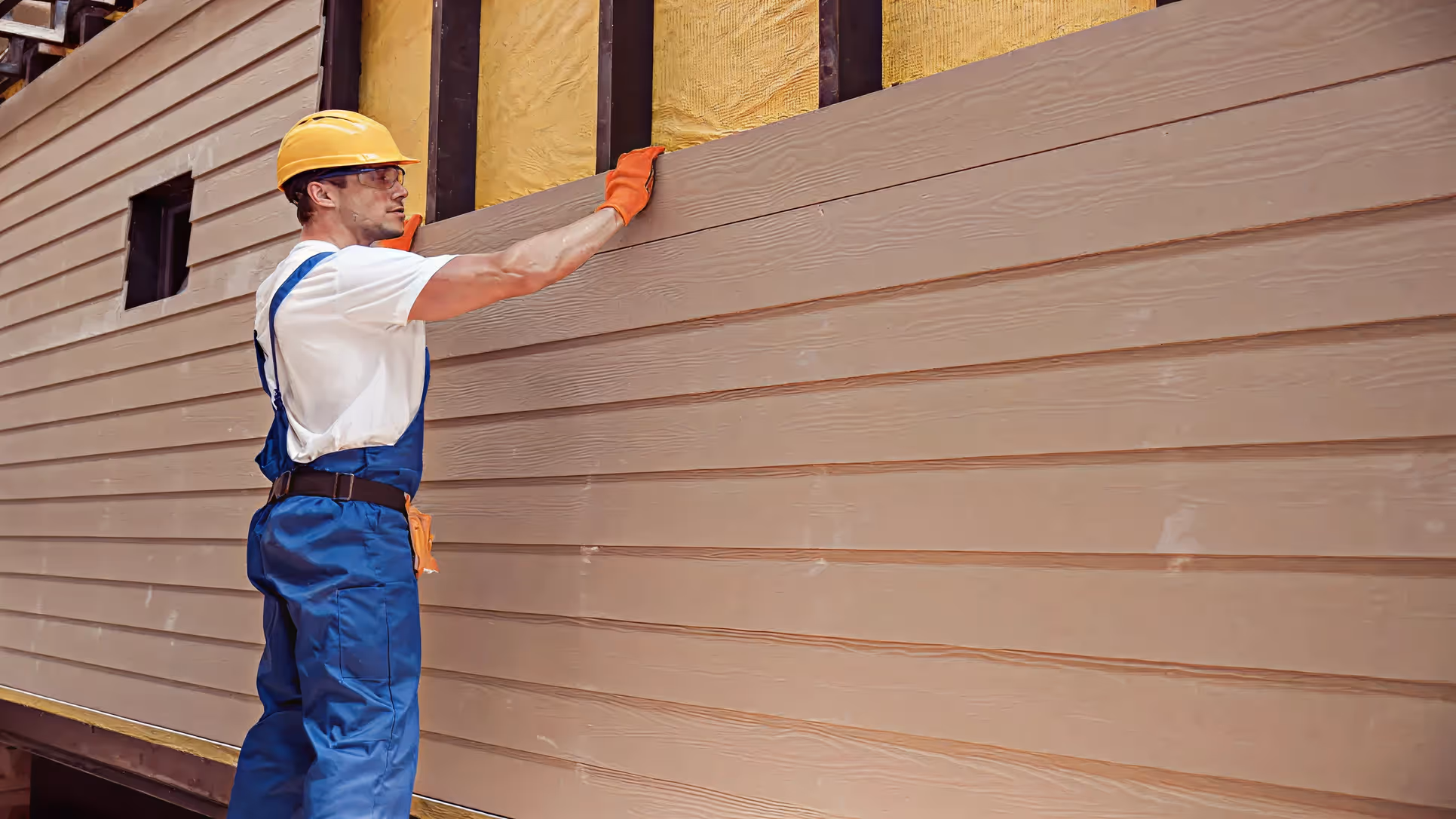 Construction worker in blue overalls and yellow hard hat installing beige horizontal siding on a building exterior.