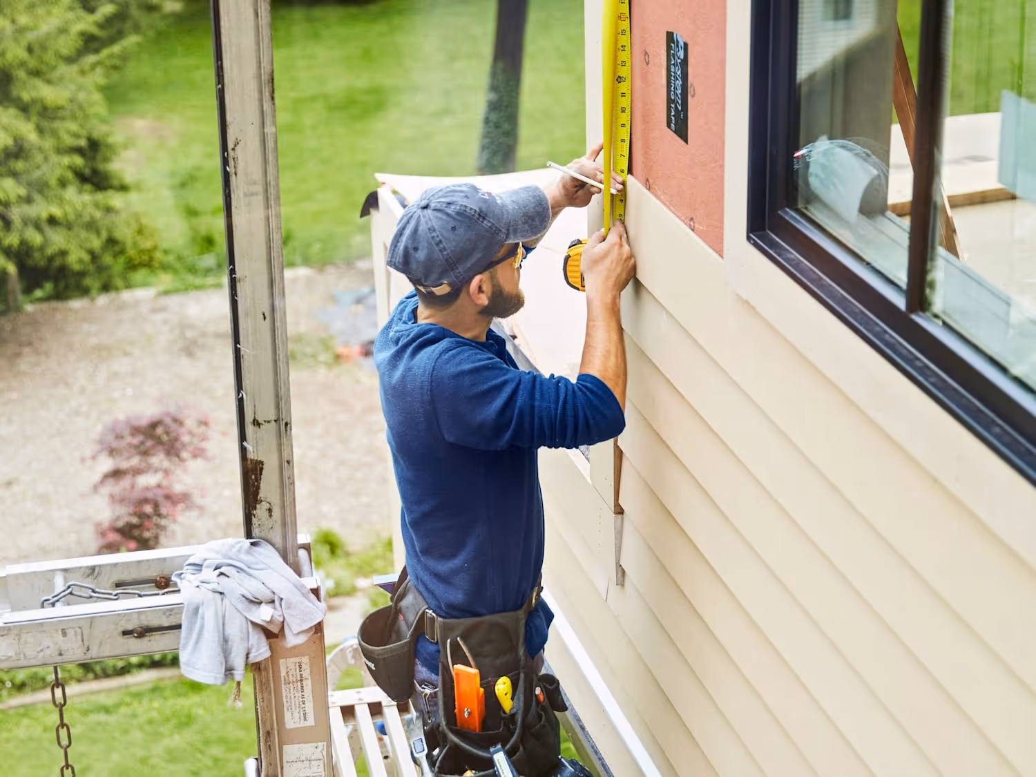 Construction worker measuring siding on a house exterior near a window, wearing a blue shirt and tool belt.