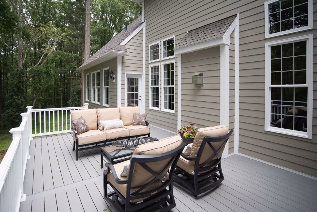 Outdoor deck with gray flooring, white railing, beige cushioned sofa and chairs, and house siding with multiple windows.