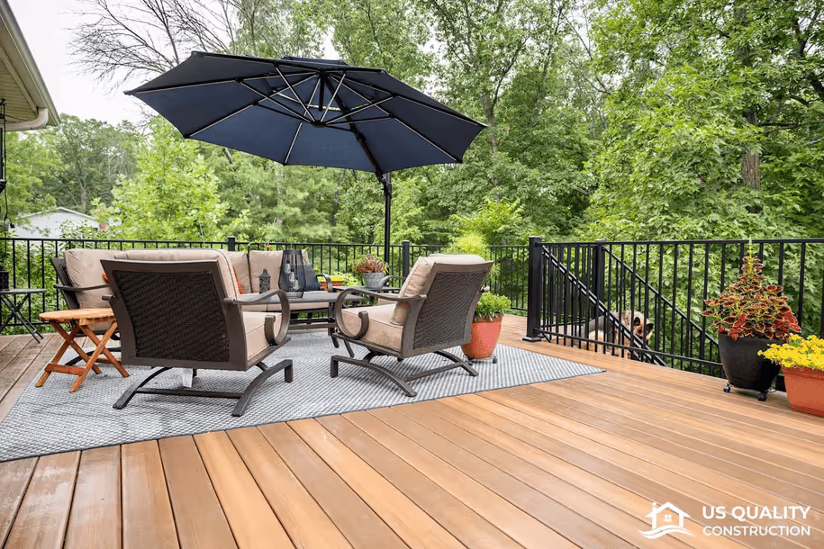 Outdoor deck with cushioned patio chairs, a table under a large navy umbrella, and potted plants surrounded by trees.