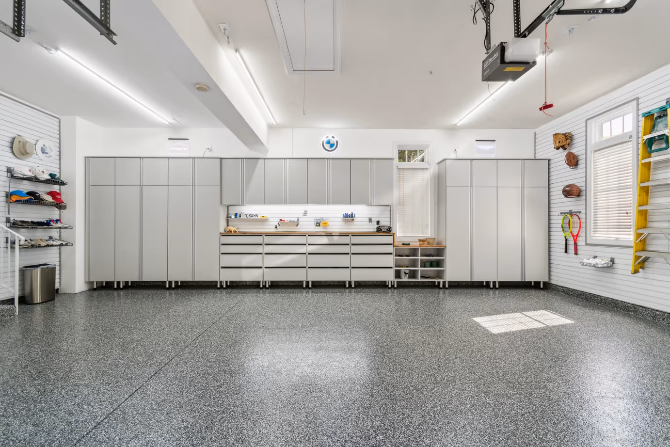 Clean and organized garage with gray storage cabinets, shelves with sports equipment, and a speckled gray floor.