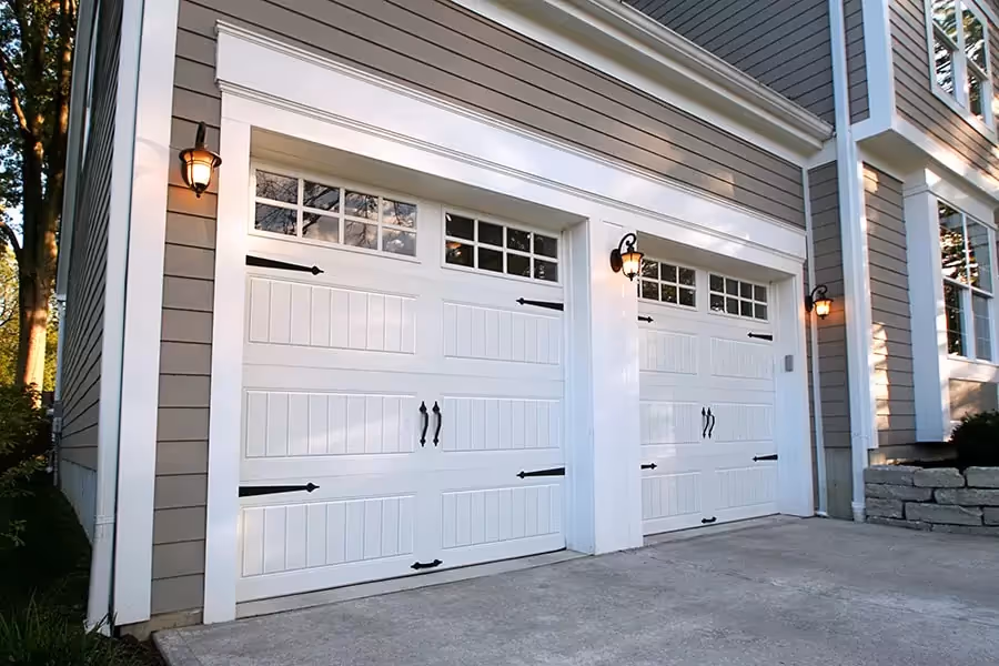 Two white paneled garage doors with black handles and hinges on a gray house exterior under warm wall lanterns.