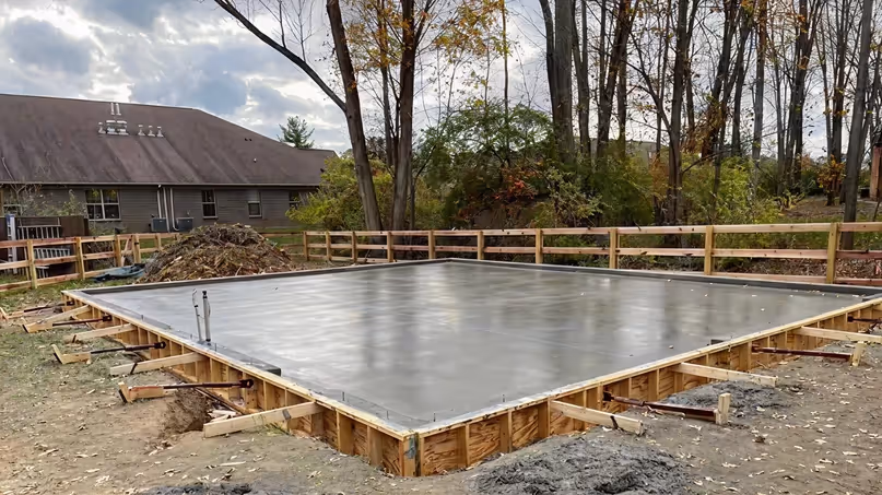 Freshly poured concrete slab foundation at a construction site surrounded by wooden framing and trees in the background.