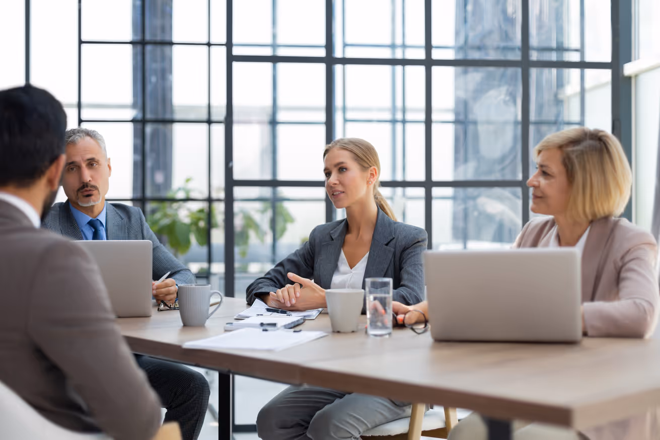 Four business professionals having a meeting around a table with laptops and documents in a modern office with large windows.