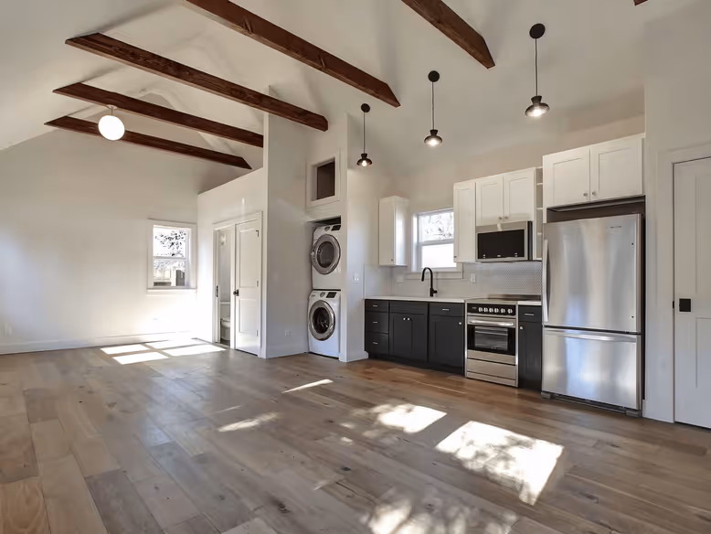 Bright open kitchen with wood floors, exposed ceiling beams, black and white cabinets, stacked washer and dryer, and stainless steel appliances.