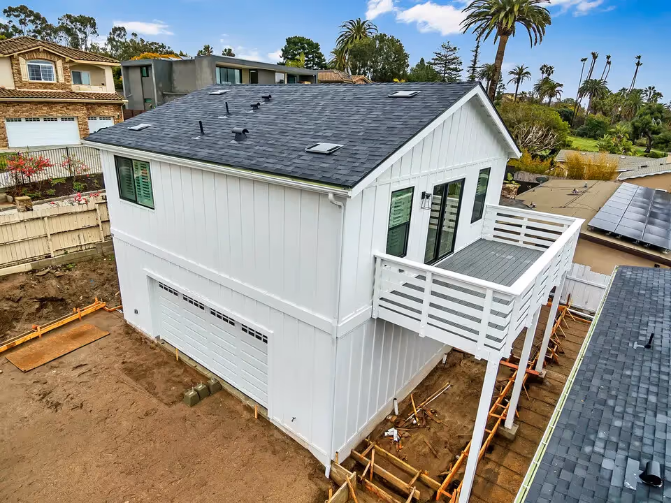 Newly constructed two-story white house with black shingle roof, balcony, and three-car garage on a dirt lot.