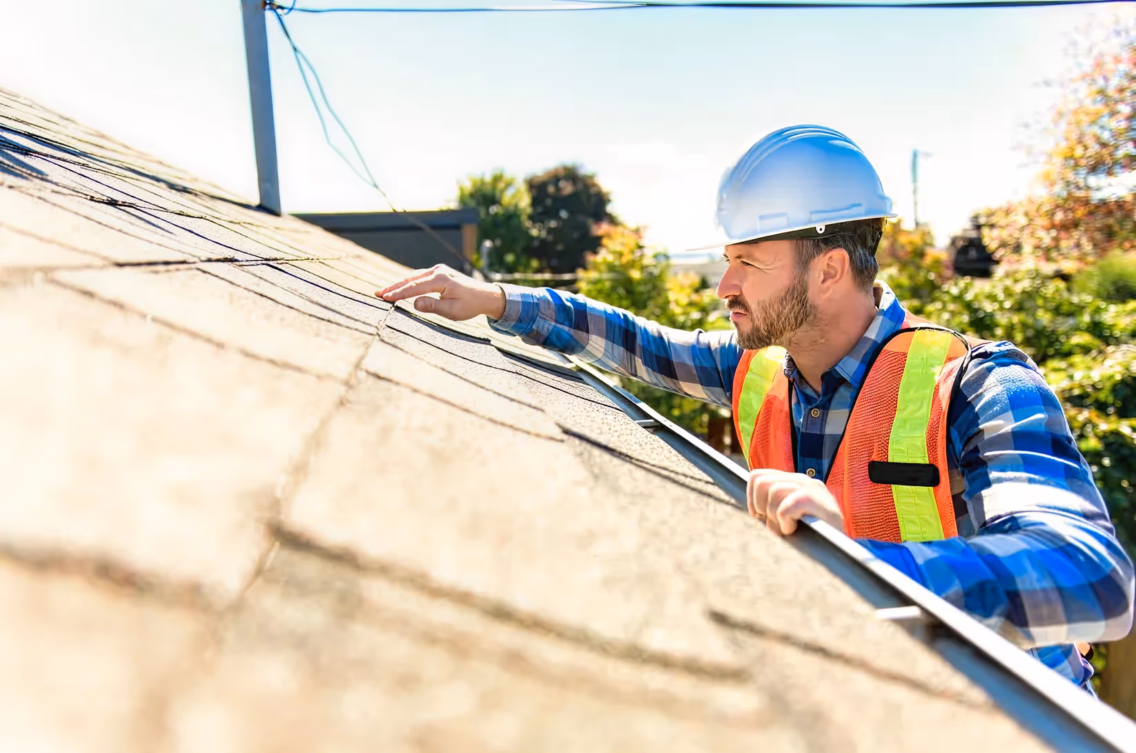 Home inspector in a hard hat and safety vest examining roof shingles closely on a sunny day.