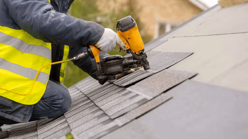 Worker in a yellow safety vest installing shingles on a roof using a pneumatic nail gun.
