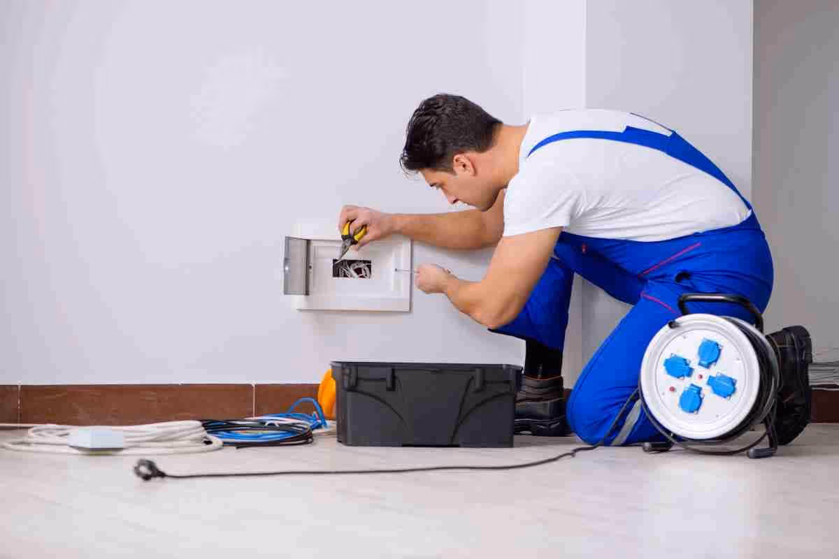 Electrician in blue coveralls kneeling and repairing wiring inside a wall outlet box.