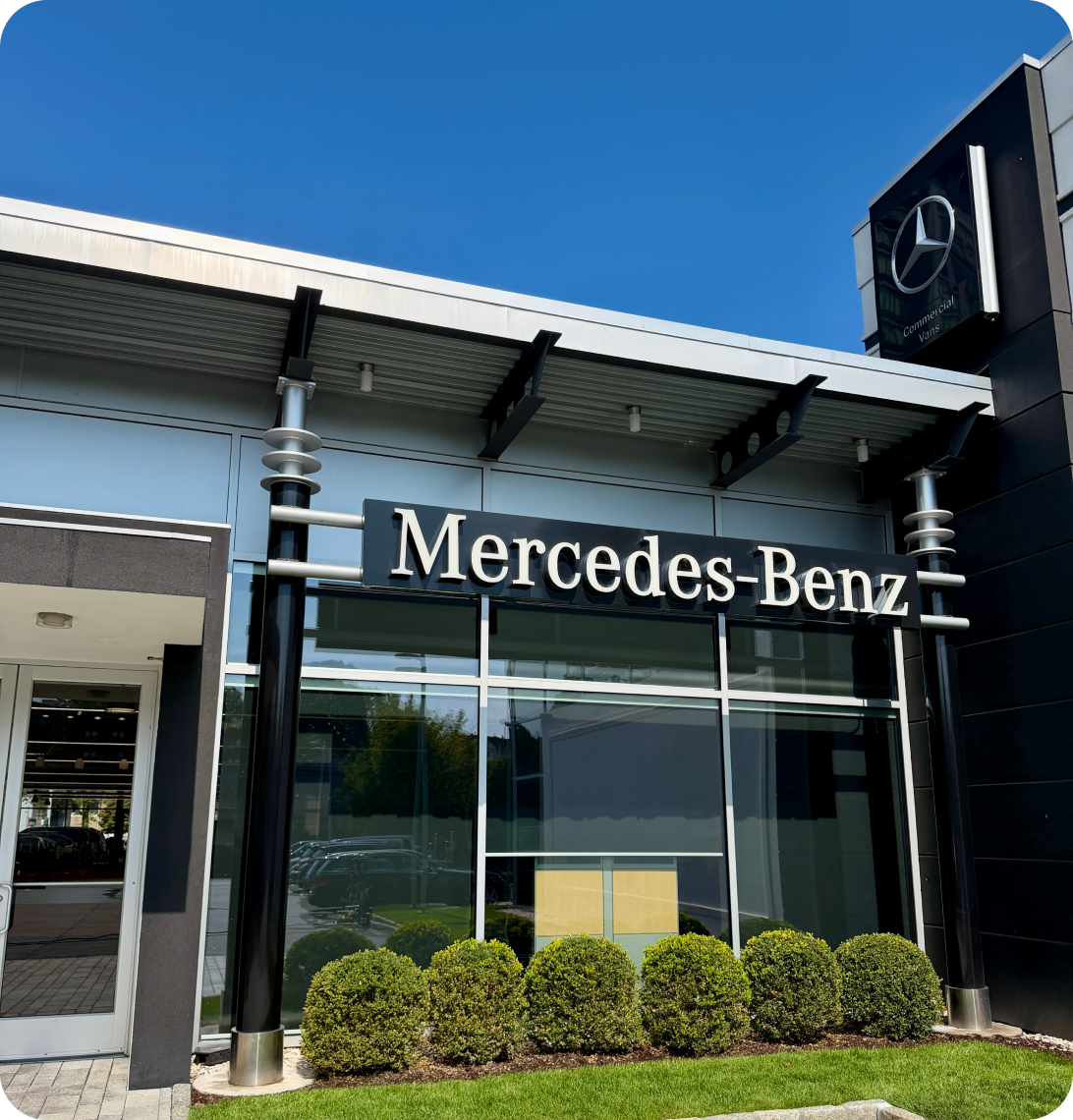 Facade of a Mercedes-Benz dealership building with the brand logo and name displayed above the entrance and neatly trimmed bushes in front.