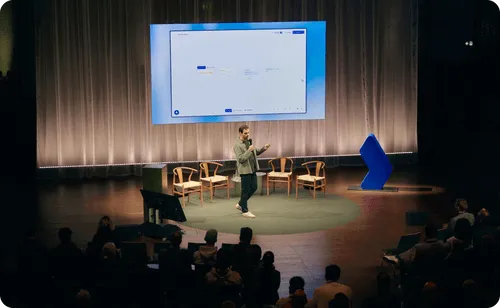 Man holding a microphone presenting on stage with chairs behind him and a large screen displaying a software interface.
