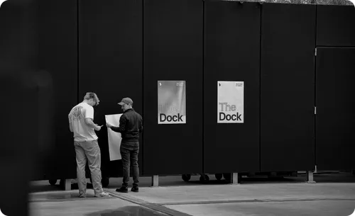 Two men standing in front of black panels with posters reading 'The Dock', one holding a large sheet of paper.