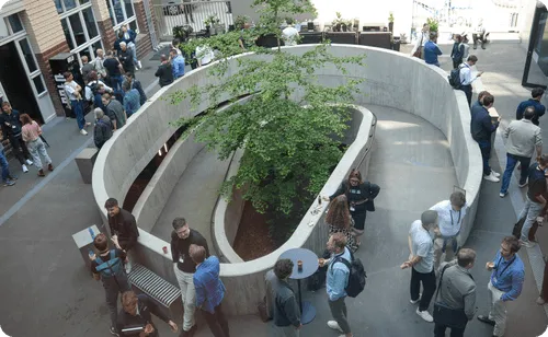 Overhead view of a modern indoor courtyard with a spiral concrete planter containing a tree, surrounded by people socializing and standing in groups.