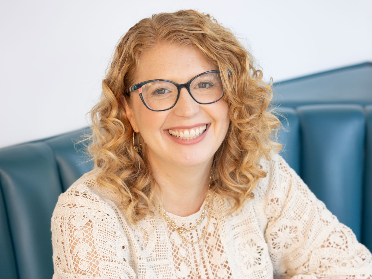 Smiling woman with curly blonde hair and glasses wearing a cream lace top, seated on a blue cushioned bench.