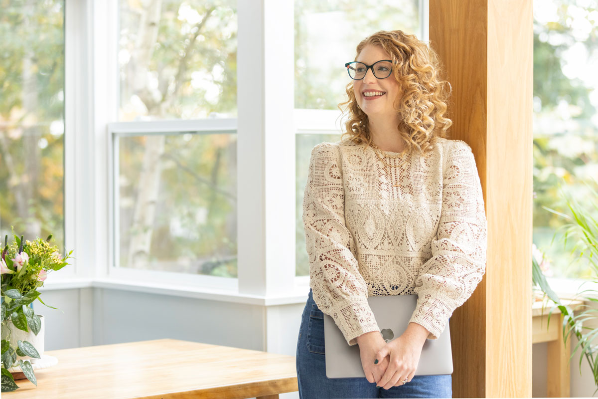 Smiling woman with curly hair and glasses holding a laptop, standing near a wooden post in a bright room with large windows and plants.