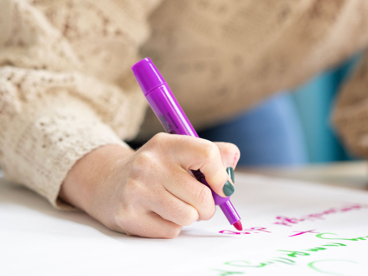 Person writing on white paper with a purple marker, wearing a beige knitted sweater.