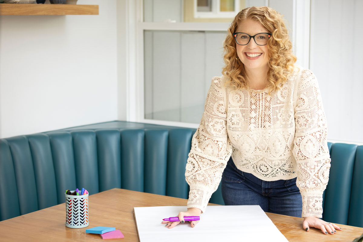 Smiling woman with curly blonde hair and glasses leans on a wooden table with markers and blank paper in a bright room.