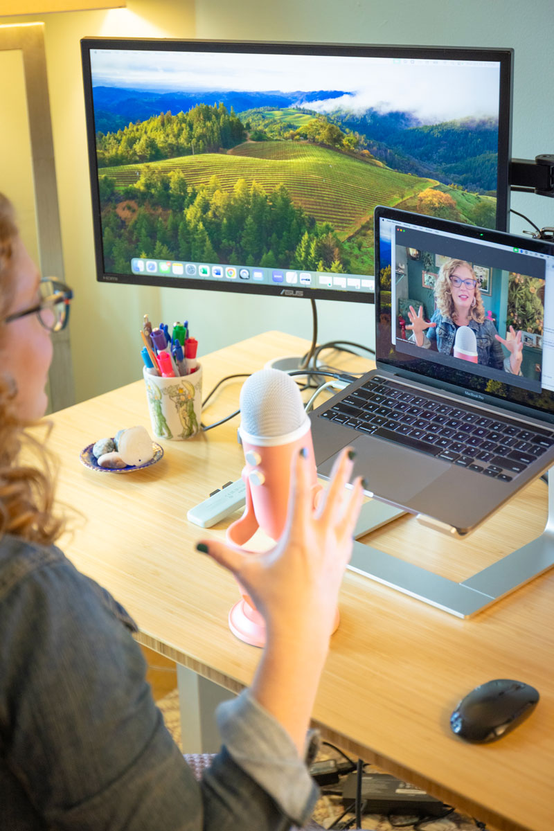 Person with curly hair and glasses waving during a video call on a laptop, with a large microphone and an external monitor displaying a scenic landscape in the background.