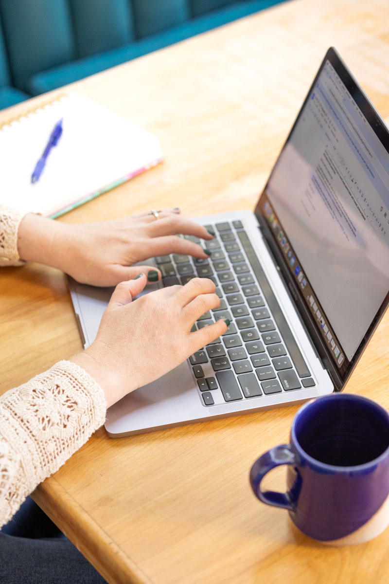 Person typing on a laptop keyboard at a wooden table with a blue coffee mug and a notebook with a pen.