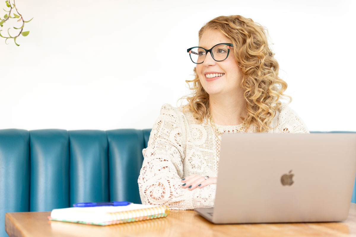 Smiling woman with curly hair and glasses sitting at a table with a laptop and notebooks.