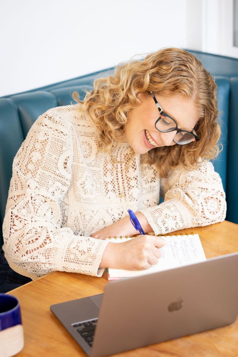 Woman with curly hair and glasses smiling while writing in a notebook at a table with a laptop.