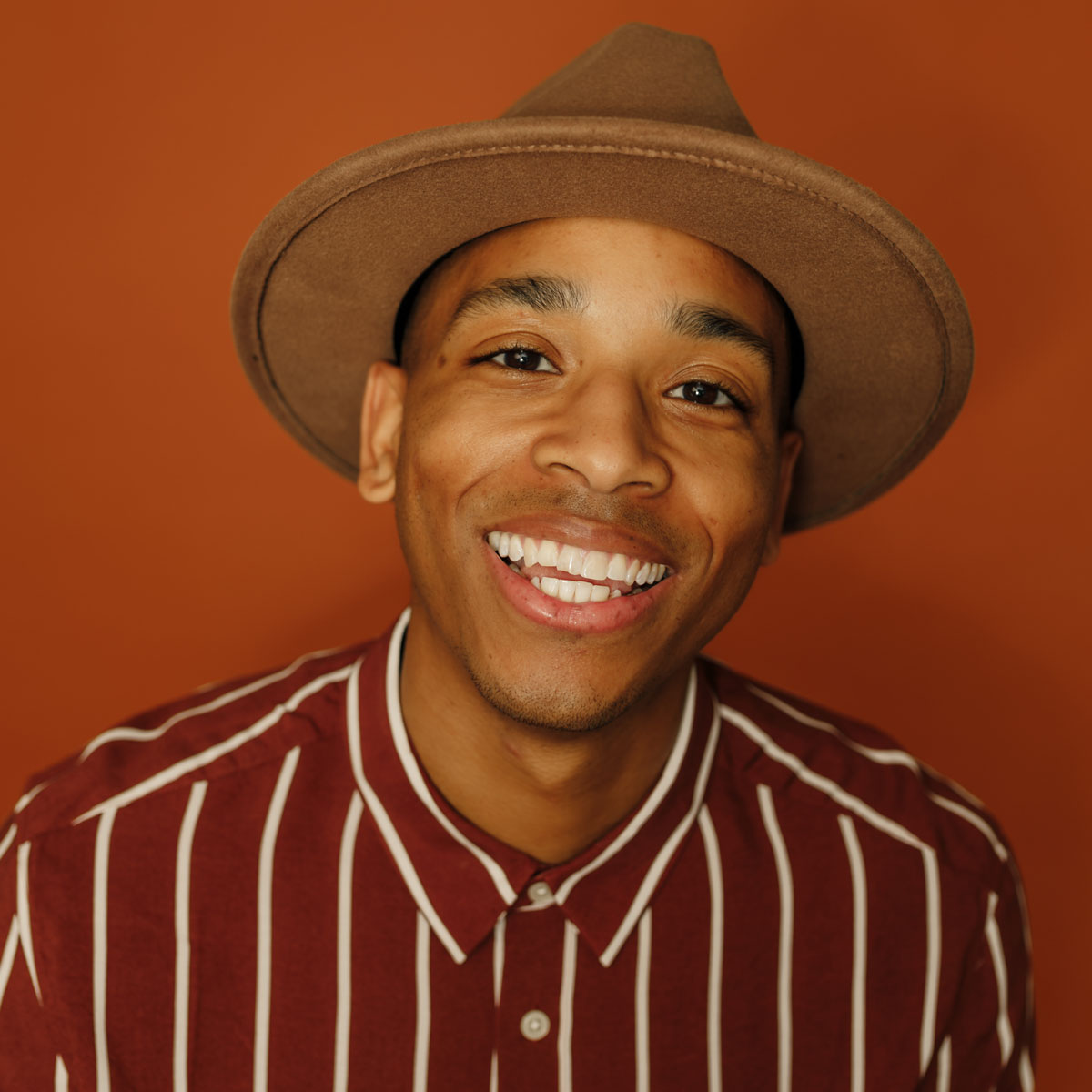 Smiling young man wearing a brown hat and red striped shirt against an orange background.
