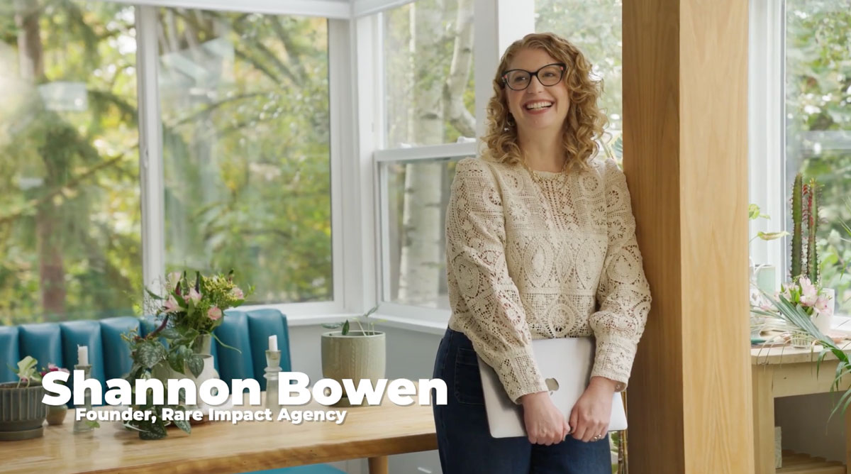 Smiling woman with curly hair and glasses holding a laptop, standing indoors next to a wooden pillar with plants and large windows in the background.