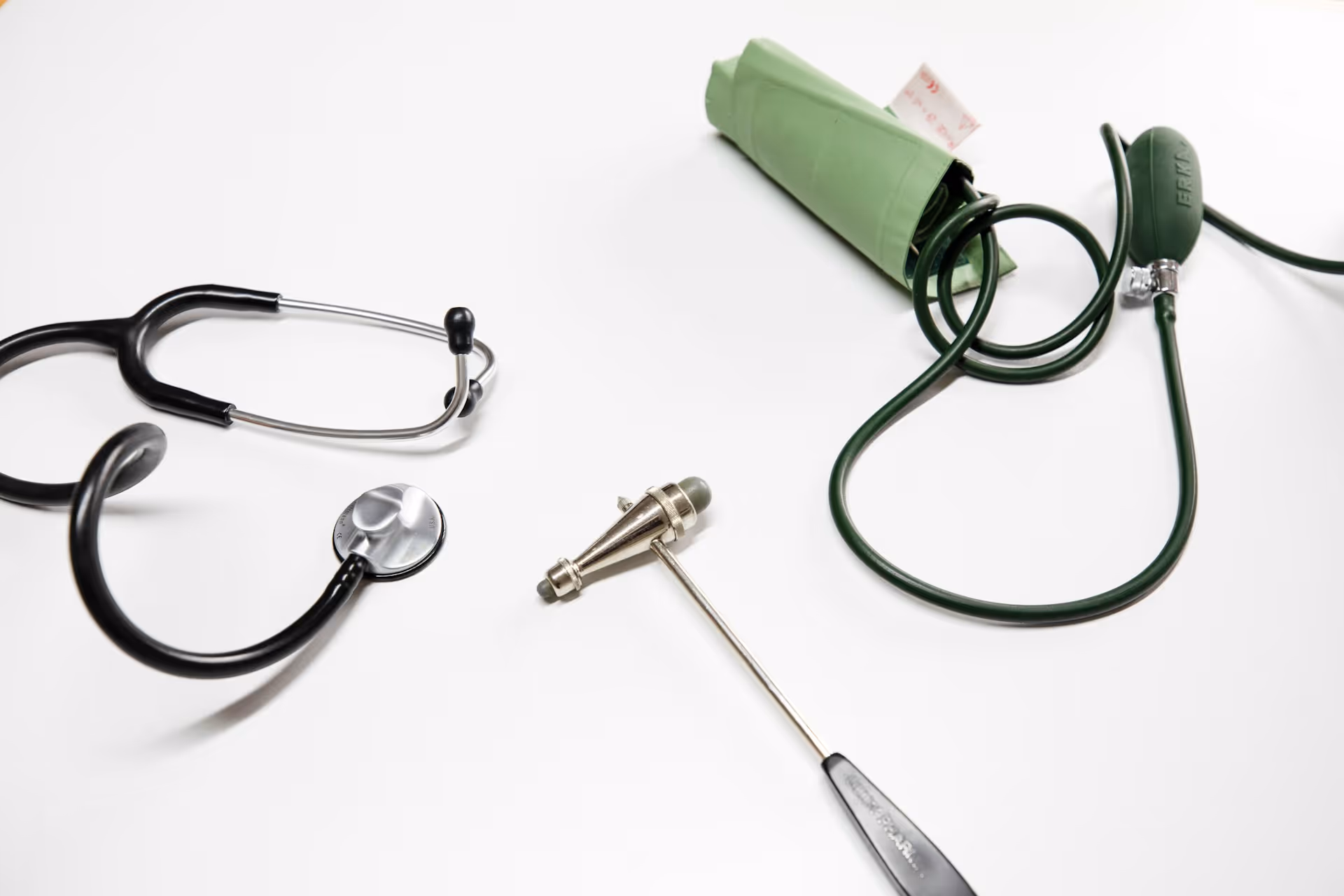 Black stethoscope, green blood pressure cuff, and reflex hammer on white surface.