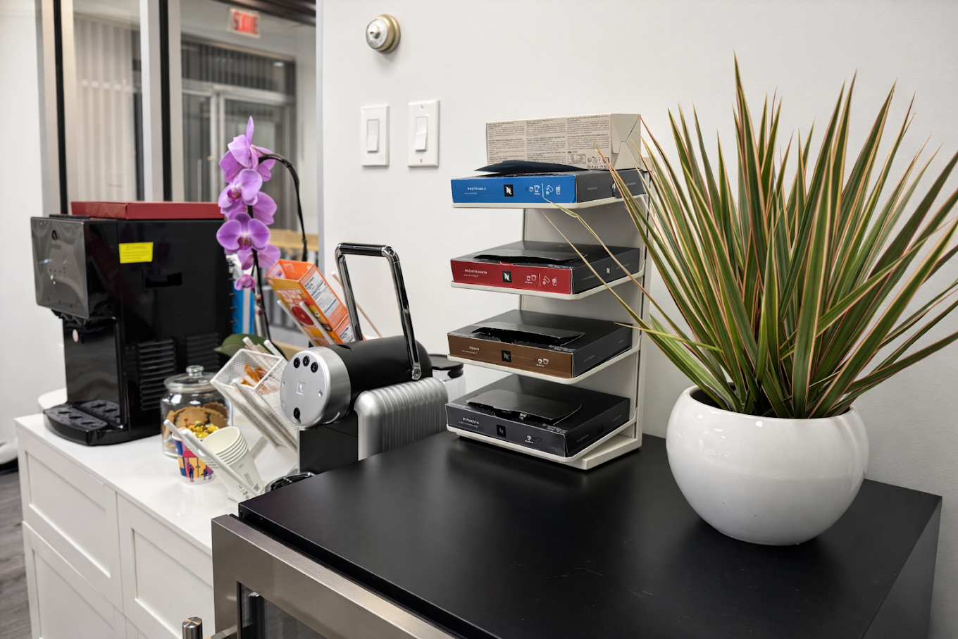 Office coffee station with a coffee machine, capsule holder with four rows of pods, orchid flower, snack containers, and a potted plant on a black countertop.