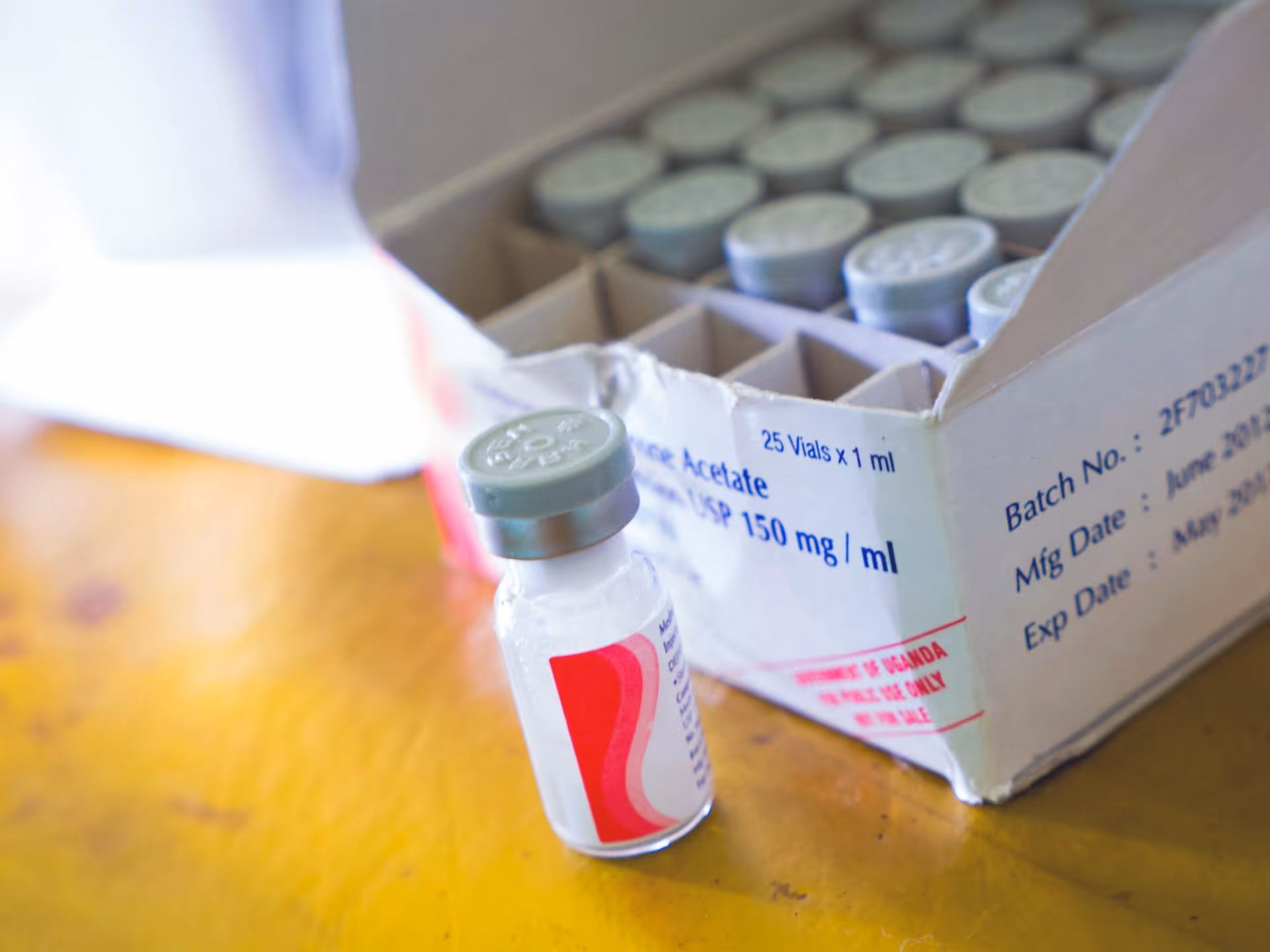 Vial of medication with gray cap in front of a box containing multiple vials on a wooden surface.