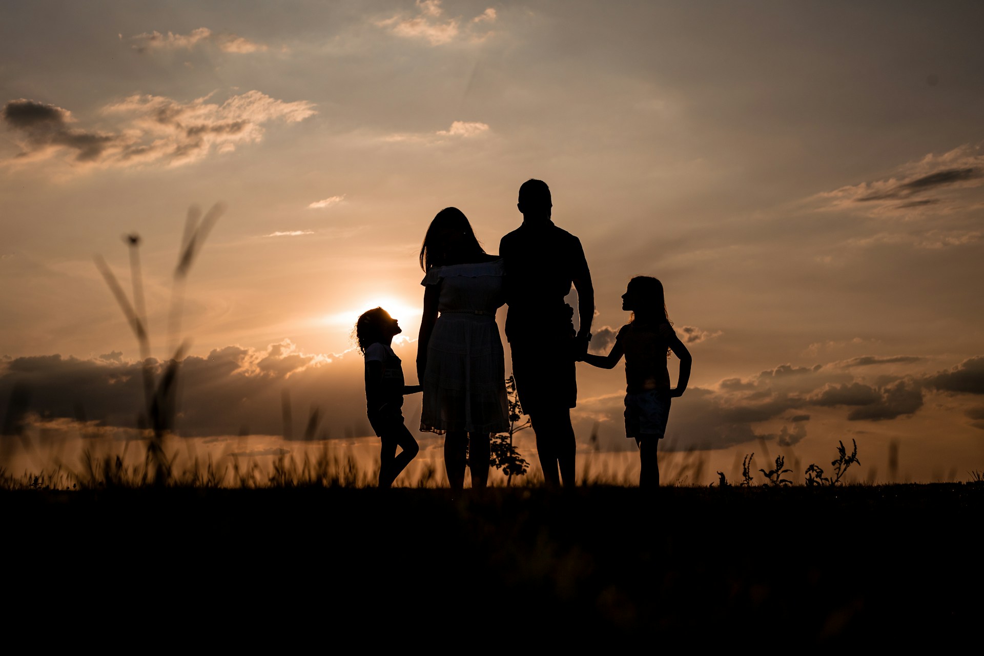 Silhouetted family of four holding hands outdoors at sunset with a partly cloudy sky.