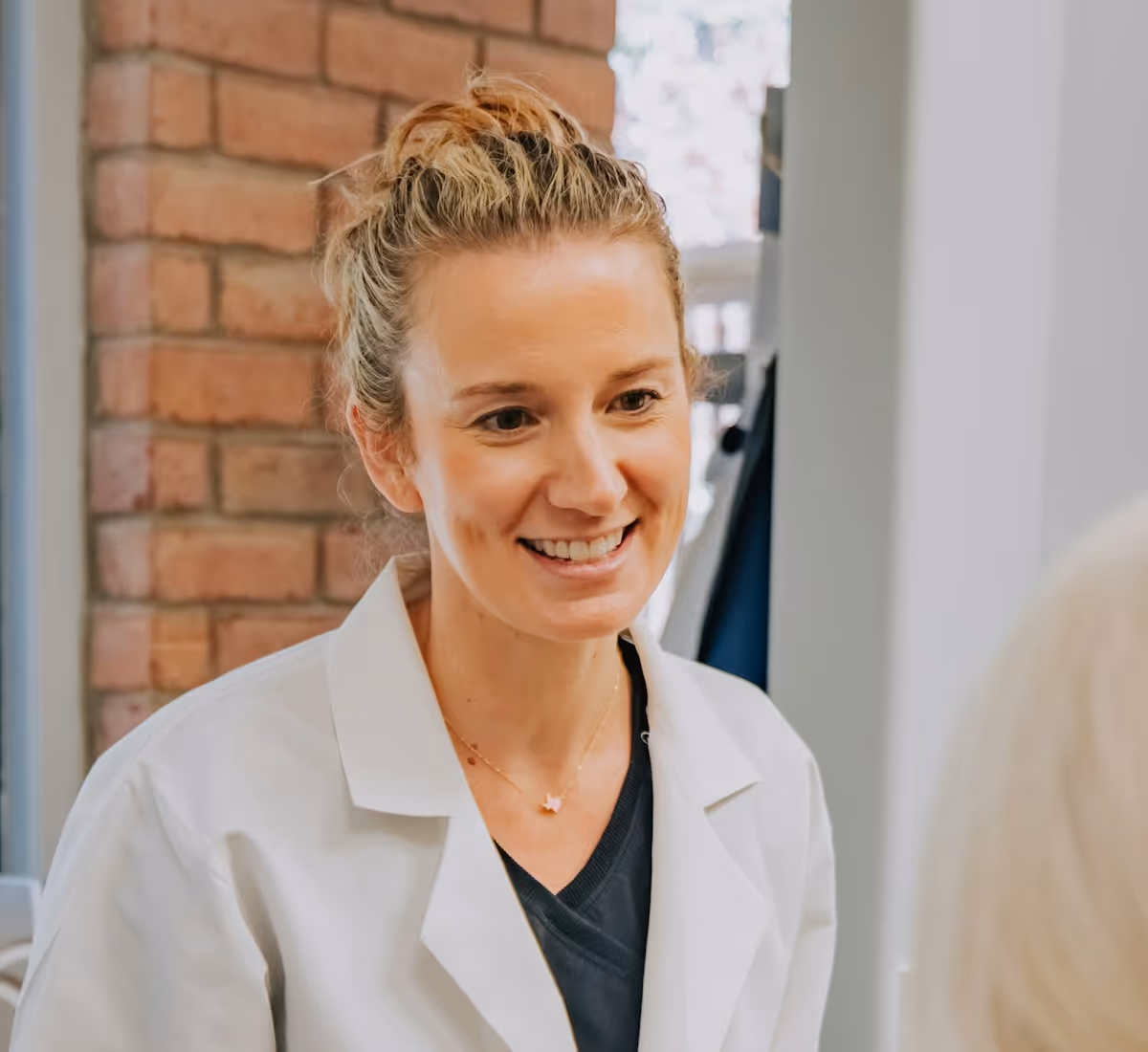 Smiling female doctor wearing a white coat and black shirt talking to a patient indoors.
