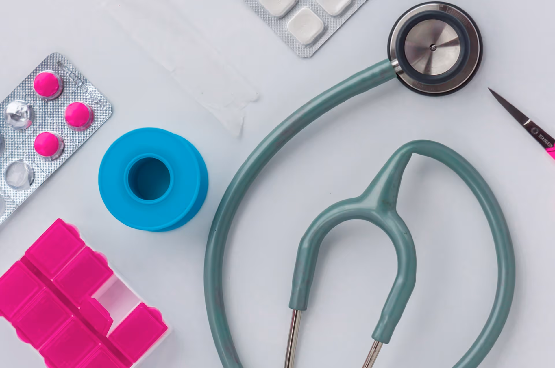 Flat lay of medical items including a stethoscope, pink and white pills in blister packs, a blue adhesive tape roll, pink pill organizer, and scissors on a white surface.